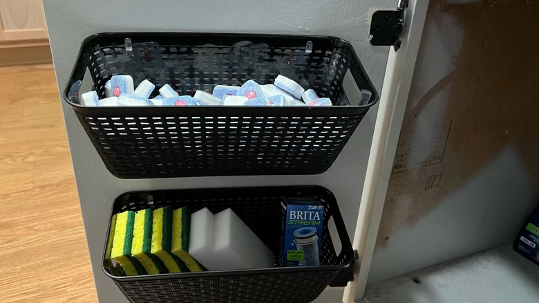 Plastic baskets with sponges and dishwasher pods hanging on inside of cabinet door via small hooks