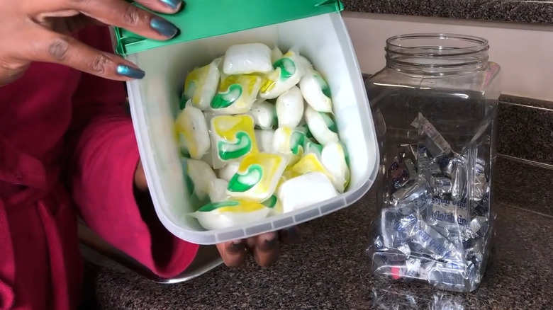 Hand with metallic green fingernails holds bin of dishwasher pods next to plastic container filled with pods on counter