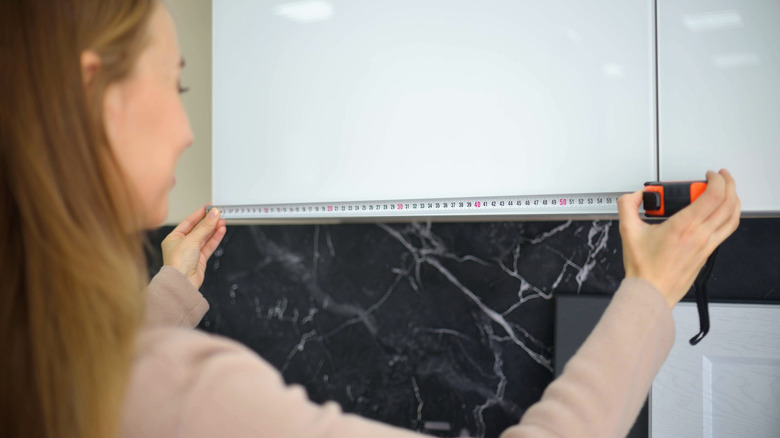 A woman in a beige sweater measuring a white cabinet with measuring tape
