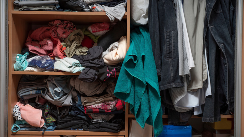 A cluttered closet with some hanging clothes and some clothes stuffed into shelves