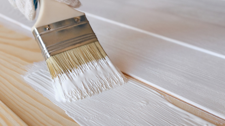paintbrush applying white paint on natural wood