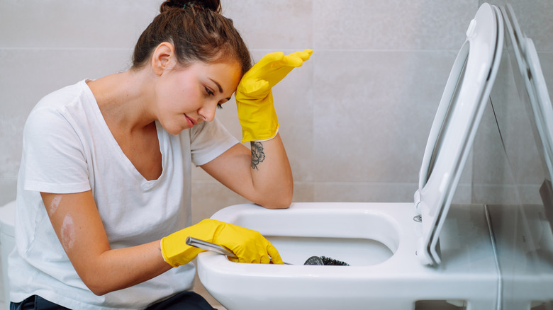 Woman toils over cleaning the toilet, she wears yellow rubber gloves and has a black scrub brush