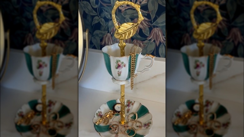 A jewelry stand is made of a vintage teacup and saucer with ornate gold cake stand hardware.