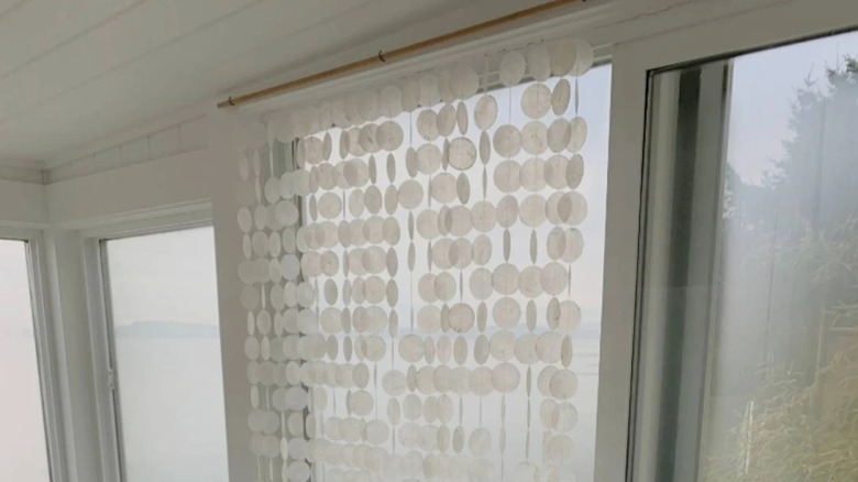 A garland made of white seashell discs hangs in front of a window in a home.