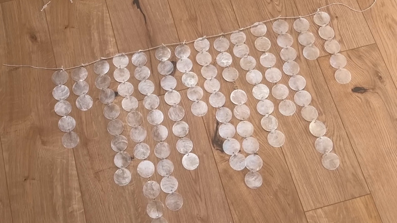 A garland made of seashell discs, jump rings, and natural cord is spread out on a wooden floor.