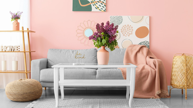 A pink room with artwork on the wall, a white coffee table, a coral blanket draped across a gray couch, shelves, and wicker decor