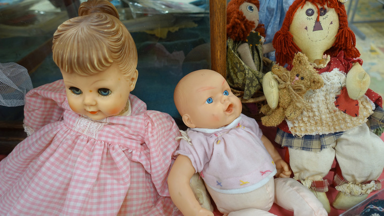 Baby doll in pink gingham dress, open-mouthed baby doll in pink shirt next to a folk doll with a bear