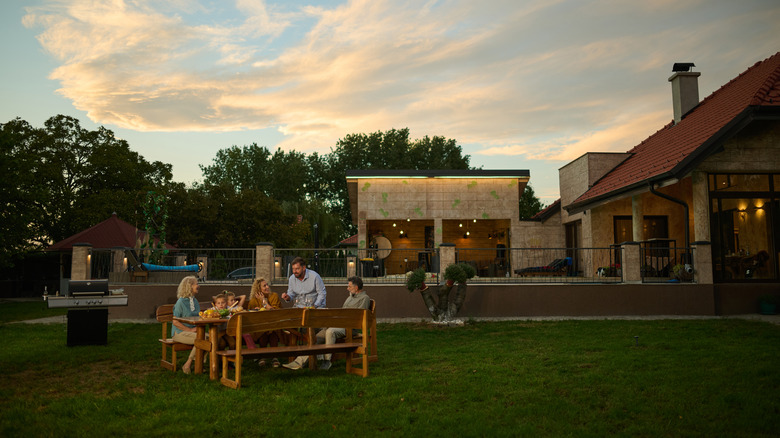 Family is gathered around a table in their backyard, enjoying a barbecue dinner party as the sun sets behind their home