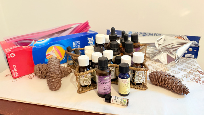 A large collection of essential oils sits on a wooden table surrounded by pinecones, a roll of aluminum foil, and a box of Ziplock gallon bags
