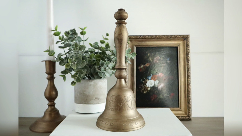 A gold dinner bell in front of a plant and framed picture.
