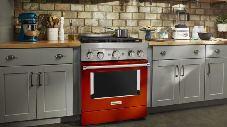 A kitchen with natural lighting, gray cabinets, wood counters, a burnt orange oven, various small appliances on the counters, and a brick backsplash.