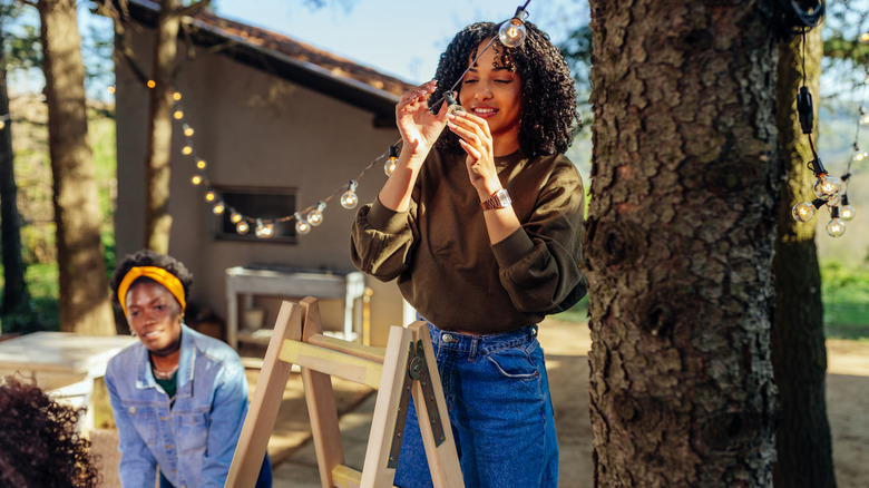 Two women hang string lights between trees in a backyard.