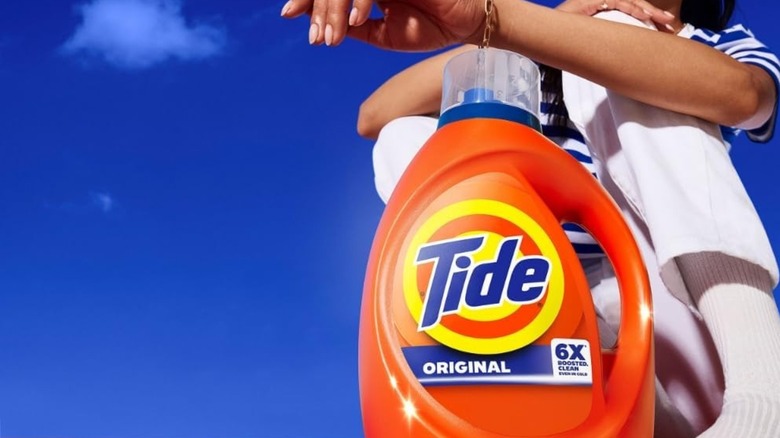 Someone crouched down next to a bottle of Tide Liquid Laundry Detergent with blue sky in the background