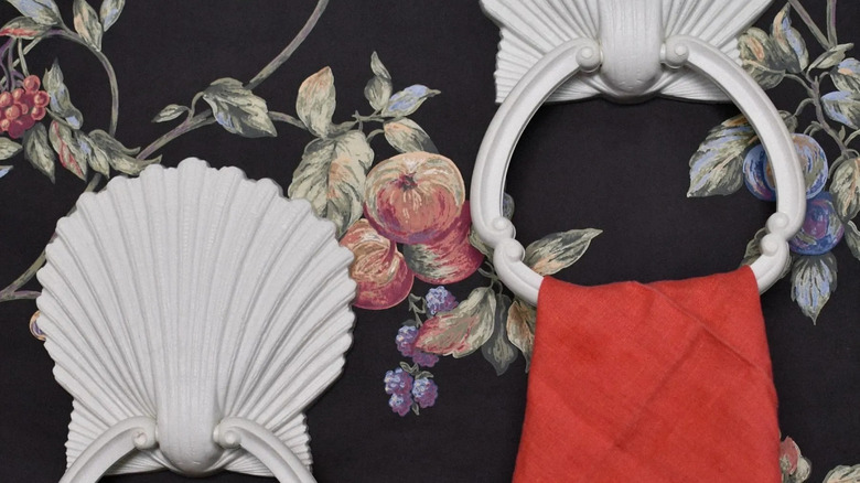 A pair of Homco shell towel rings with a red hand towel hanging off one of them are mounted to a wall with floral wallpaper.