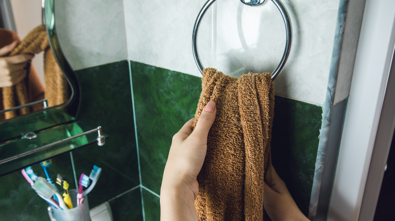 A person dries their hands on a brown towel hanging from a simple towel ring mounted to a tiled bathroom wall.
