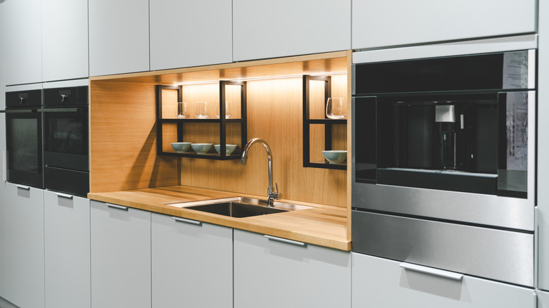 A silver kitchen sink, faucet, and appliances set into a wood countertop and backsplash and gray kitchen cabinets.