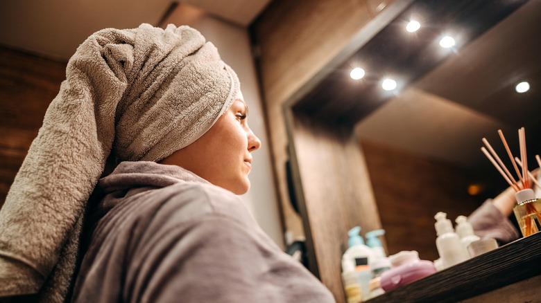 A young woman in a robe with a towel on her head gets ready in front of a bathroom mirror.