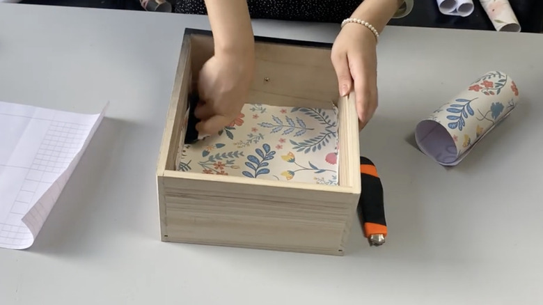 A pair of arms laying down flower-themed wallpaper in a wooden drawer.