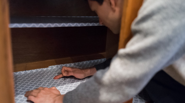 A man sticking his upper body into a two-shelved cabinet is putting down a gray and white liner.