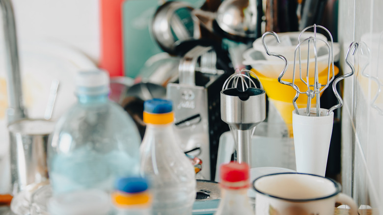 A cluttered kitchen bench with small appliances, bottles, and a coffee mug