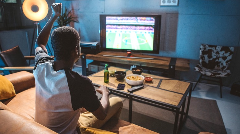 Man on leather sofa eating snacks and watching soccer in man cave