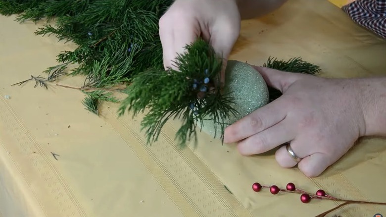 Person constructing kissing ball for Christmas by putting greenery clippings into Styrofoam ball