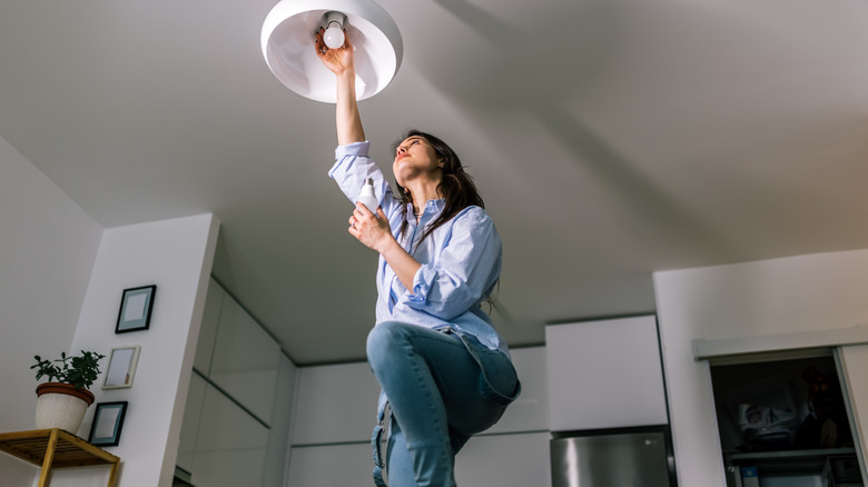 Woman standing on ladder installing light bulb in white light fixture