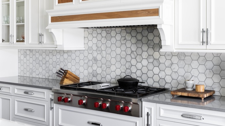 A kitchen range detail with a marble hexagon tile backsplash, white cabinets, stainless steel stovetop and range hood, and gray countertop.
