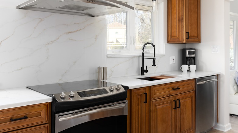 A kitchen detail with a stainless steel stove and range hood, a white calacatta marble slab backsplash and white countertop, wood cabinets, and a gold and black light hanging above the black faucet.