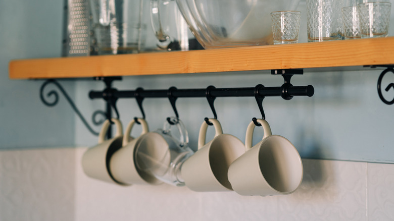 A set of grey mugs hanging off small black hooks installed under a wooden kitchen shelf