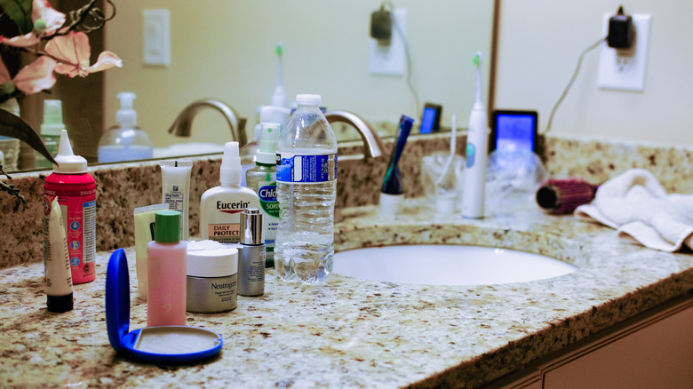 A messy bathroom vanity full of beauty products, a nearly empty plastic water bottle, an electric toothbrush, and more