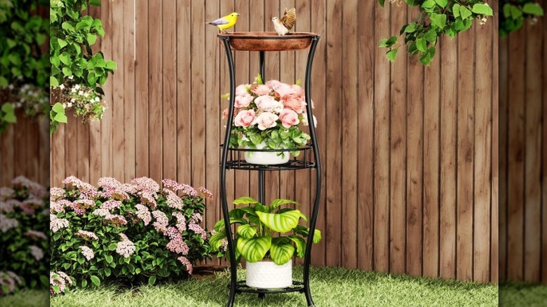 The bird bath plant stand sitting outside on the grass next to a fence with three birds enjoying the bath and some plants on the lower tiers