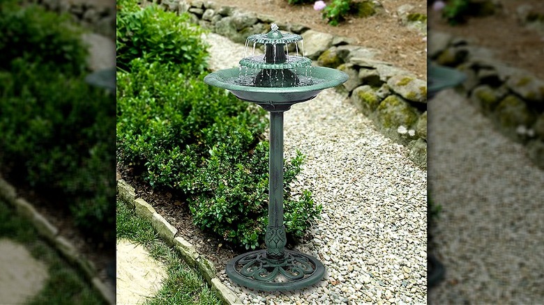 A bird bath styled to look like a vintage fountain with a green patina, positioned by a gravel path and shrubbery