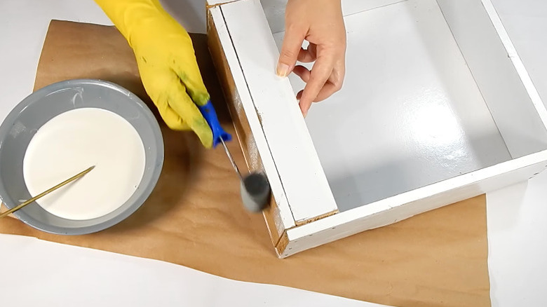 A person in one yellow rubber glove painting the edges of a drawer white
