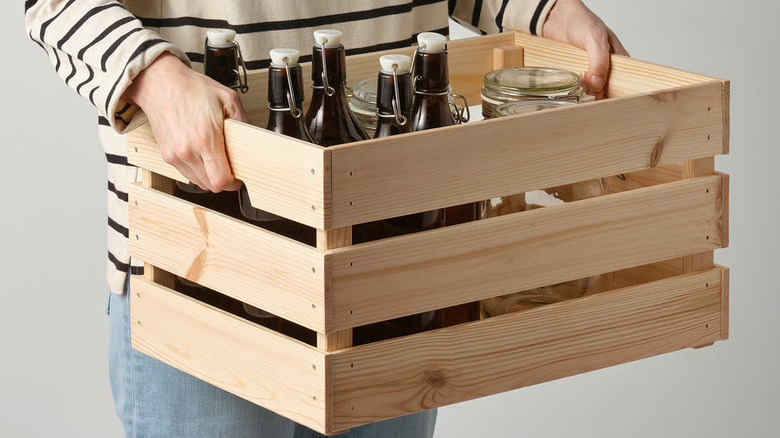 Woman holding one of the KNAGGLIG pine boxes with bottles in it