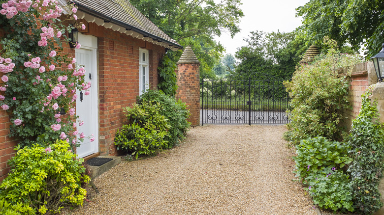 A one-story brick house surrounded by bushes and flowers with a brown gravel driveway out front leading to an iron gate
