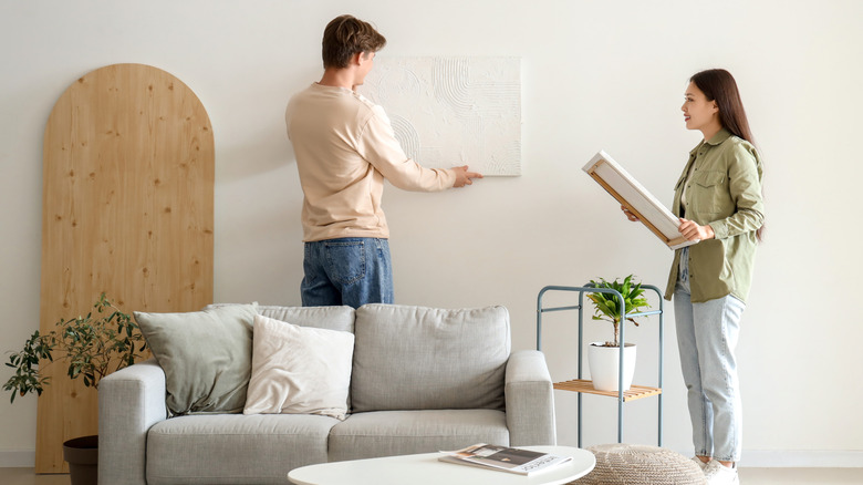 Two people hanging art on the wall behind a sofa