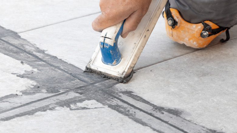 Craftsman filling tile joints with grout using a float and sitting on his knee, hand closeup detail.