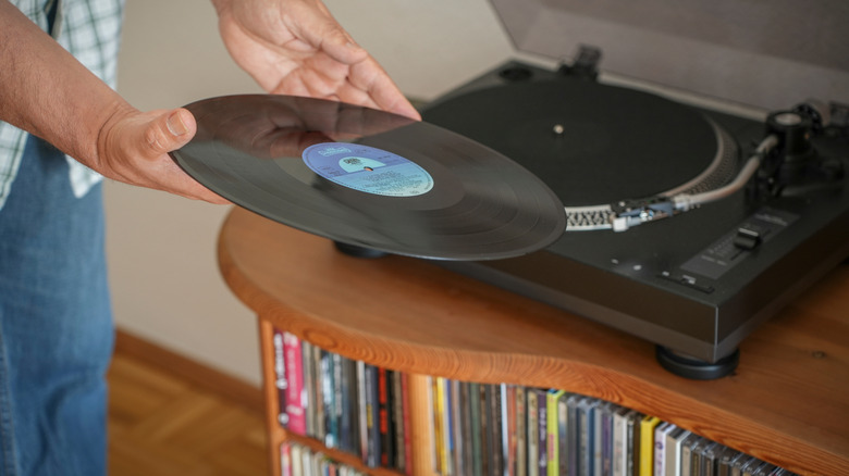 Hands loading a record onto a record player sitting on a cabinet filled with CDs.