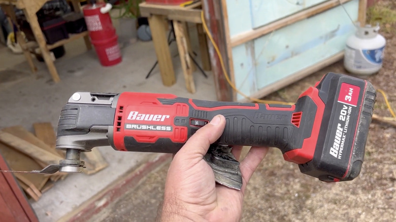 A hand holding the Bauer 20V Brushless Cordless Variable-Speed Oscillating Multi-Tool with an unkempt workstation in the background.