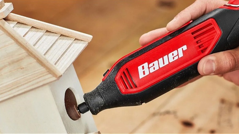 Hand using a Bauer rotary tool to work on crafting a wooden birdhouse.