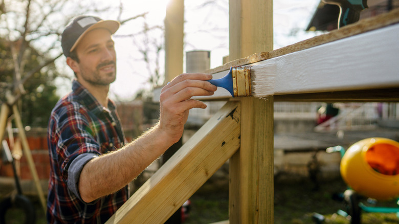 Man applying white paint to a wooden outdoor structure.