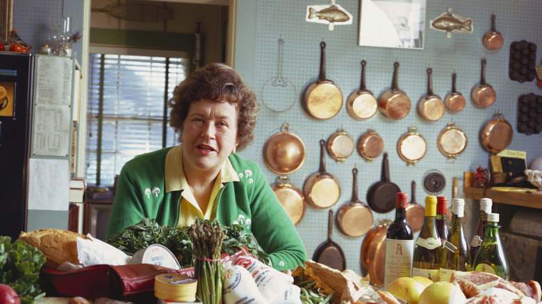 Julia Child sitting at her kitchen table surrounded by vegetables and bottles of wine.
