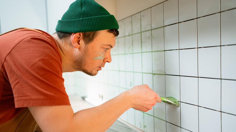 Person wearing a green hat and red shirt adds grout to white tile backsplash.