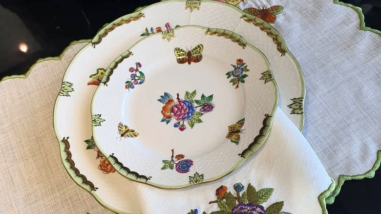 Two Herand china plates in the Queen Victoria colorful butterfly enamel pattern stacked on top of one another over a linen placemat with green borders on a black glass table.