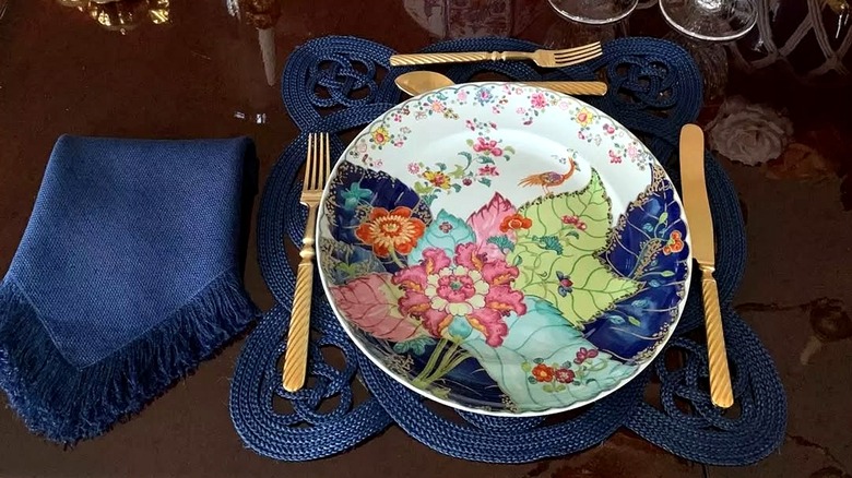 Porcelain plate in the Famille Rose Tobacco Leaf pattern with colorful tobacco leaves pink, blue, and green enamels, over a dark blue woven placemat on a glass table.