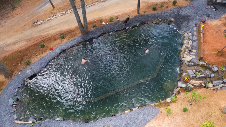 Two people swimming in a large natural pool surrounded by dirt and dirt roads