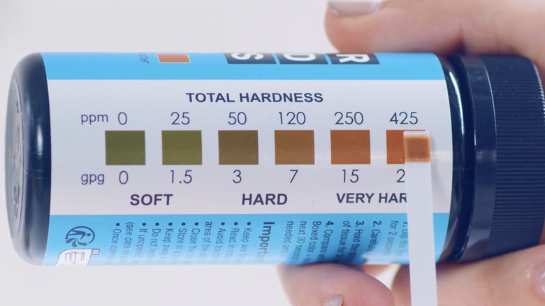 A person holds a water test strip next to a color chart on a small bottle