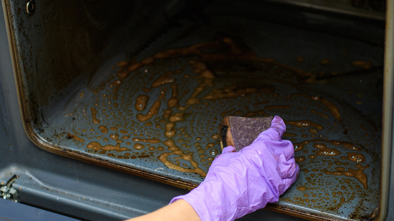 A purple-gloved hand cleans the bottom of a dirty oven with a scrubbie.