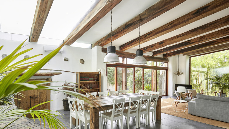 A bright room with dark ceiling beams, large windows, and a farmhouse style dining room table.